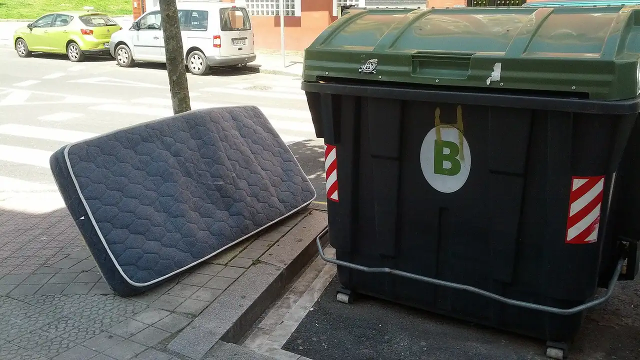 A discarded single mattress leaning against the curb beside a large street waste container