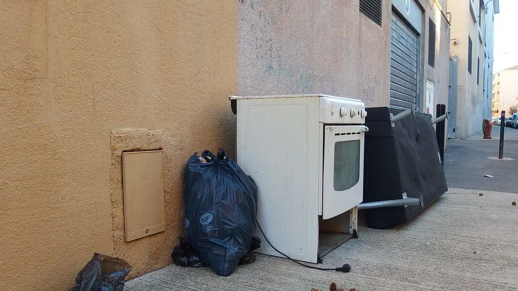 An old white stove and a full black garbage bag set against a building wall on a sidewalk